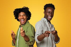 Gotcha. Two Cheerful African American Guys Poiting Fingers At Camera, Joyful Young Black Male Friends Saying Hey You, Laughing And Having Fun While Posing Over Beige Studio Background, Copy Space photo