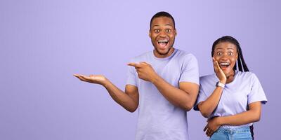 Omg, Check This. Portrait Of Emotional Black Man Pointing Finger Aside At Copy Space On Open Palm Over Purple Background, Surprised Woman Looking At Camera, Demonstrating Free Place For Your Advert photo