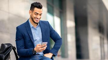 Wealthy middle-eastern entrepreneur using cellphone, sitting on the street by office building with backpack and laptop, resting after business event, checking emails, using business app, copy space photo
