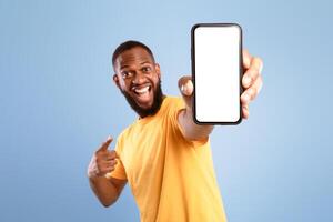 Mobile app advertisement. Excited African American man pointing at smartphone with empty screen on blue studio background, mockup. Cellphone display template, space for ad or website photo