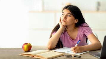 Thoughtful Indian teen girl studying online from home, taking notes during web lesson, feeling bored at home. Pensive adolescent participating in dull lecture, learning remotely during covid lockdown photo
