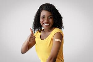 Covid-19 Vaccination Concept. Vaccinated African American Lady Gesturing Thumbs Up Showing Arm With Medical Plaster After Coronavirus Vaccine Injection Sitting In Studio Over Gray Background photo