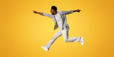 Crazy Sales. Excited black guy jumping up in air and pointing aside at copy space on beige background, emotional young african man demonstrating free place for design or advertisement, panorama photo