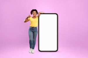 Advertisement for mobile application. Cheery African American lady standing near modern smartphone with empty screen and showing thumb up gesture on violet studio background, mockup photo