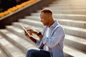 Millennial black guy browsing internet on tablet computer while sitting on stairs outside photo