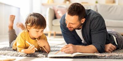 Kids development and art hobby concept. Young loving father drawing with his little son, painting in album with pencils, lying on floor at home, panorama photo