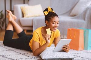 Online Transaction Concept. Smiling black lady using digital tablet and holding credit card, ordering purchases via internet, lying on the floor carpet with pillow and shopping bags photo