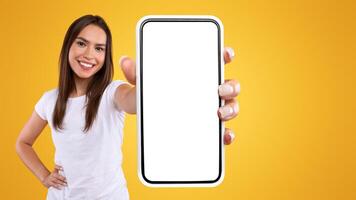 App Ad. Smiling pretty woman holding smartphone with white blank device screen in hand close up to camera, orange studio. Gadget with empty free space for mockup, banner isolated, selective focus photo