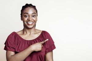 Pretty african american young lady in casual outfit posing alone on beige studio background, pointing at copy space and smiling, panorama. Black woman showing empty space for text or advertisement photo