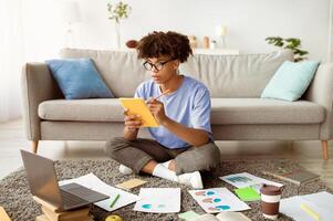 Focused black teen student with laptop taking notes for exam during online studies at home, full length. Smart teenage guy working on assignment or coursework paper, learning remotely photo