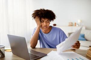 Portrait of bored black teen sitting at desk with documents, using laptop to write coursework paper, studying online, making difficult college home assigment, preparing for web exam, indoors photo