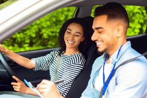 Cheerful middle-eastern guy instructor examinating happy brunette lady student, taking notes at chart while sitting by young woman driving car and waiting for test results, side view, copy space photo