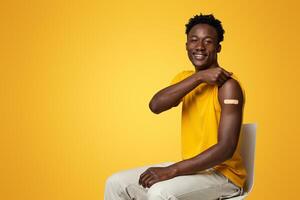 Im Vaccinated. Black Guy Showing Arm With Adhesive Bandage After Successful Vaccination, Young African American Man Got Injection Shot Against Covid-19, Sitting On Chair Over Beige Background photo