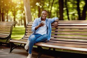 Smiling black man with mobile phone relaxing on bench at park on beautiful summer day photo