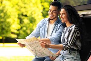 Happy young lovers sitting on auto trunk and watching road map, copy space. Handsome arab guy bonding with his beautiful girlfriend, having break while car trip, looking for destination to go photo