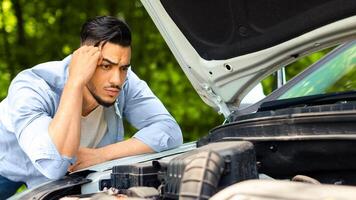 Puzzled arab man leaning on open car hood, looking at engine, having automobile crush in the middle of the road. Nervous middle-eastern guy driver waiting for car service, closeup photo