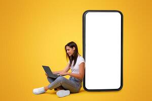 People And Technology. Smiling Woman Using And Looking At Pc Screen, Sitting And Leaning On Big Smartphone With White Blank Empty Screen For Mock Up, Isolated Yellow Studio Background, Free Copy Space photo