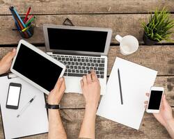 Hands interacting with various devices on a wooden table during a working session in a cozy workspace photo