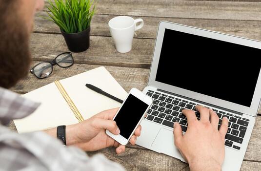 Young man working on laptop while using smartphone at rustic wooden desk in a casual setting photo