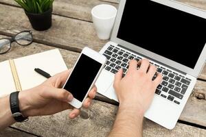 Hands using smartphone and laptop on wooden table in a casual workspace setting photo