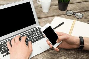 Hands typing on a laptop while holding a smartphone in a workspace setting with a cup and stationary visible photo