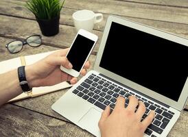 Hands typing on a laptop while holding a smartphone with a coffee cup and notepad nearby in an office setting photo