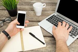 Remote work setup with smartphone and laptop on wooden desk featuring notebook and coffee cup photo