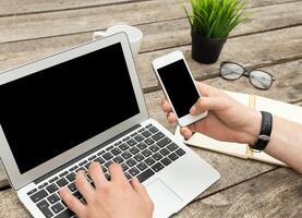 Person working on laptop while using smartphone at wooden desk with plant and glasses in a cozy office setting photo
