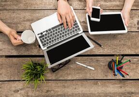 Hands engaged with various devices and a coffee cup on a rustic wooden table during a collaborative work session photo