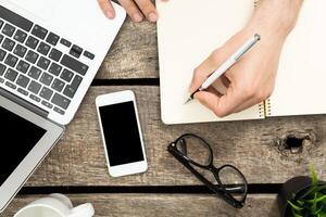 Person writing notes while using a laptop and mobile phone on a rustic wooden table during a work session photo