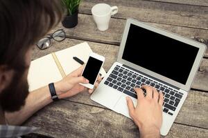 Individual using a smartphone while working on a laptop at a wooden desk in a cozy workspace photo