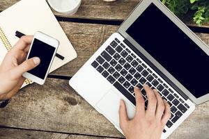 Working on a laptop while holding a smartphone in a cozy workspace surrounded by a rustic wooden table photo