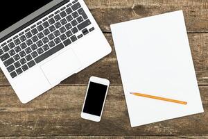 Workspace setup featuring a laptop, smartphone, blank paper, and pencil on a rustic wooden table in a cozy environment photo