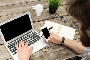Young adult engaged in multitasking with laptop and smartphone at rustic workspace during daytime photo