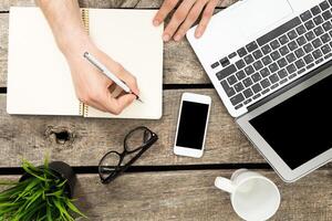 Writing notes while working on a laptop with a smartphone and coffee on a wooden table during daylight hours photo