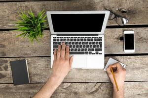 Writing notes while working on a laptop at a wooden desk with office supplies during the day photo
