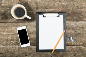 Workspace with clipboard, coffee, smartphone, and pencil on rustic wooden table during morning hours photo