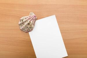 Shell rests next to blank sheet of paper on wooden surface creating a minimalistic aesthetic photo