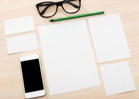Workspace setup on a wooden desk with empty paper, glasses, and a smartphone in a well-lit environment photo