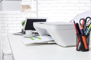 Office workspace with printer, laptop, and coffee cup on a white desk during a bright afternoon photo