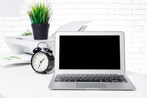 Modern workspace with a laptop, clock, printer, and greenery during daylight hours photo
