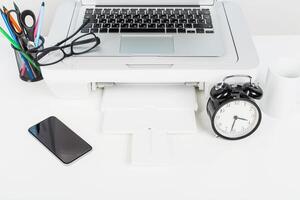 Home office workspace featuring a laptop, printer, clock, and smartphone during a bright afternoon photo