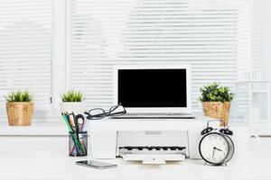 Modern workspace setup with laptop, printer, and office supplies in a bright room during the daytime photo
