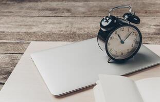 Desk arrangement featuring an alarm clock beside a laptop and an open notebook in a rustic setting photo
