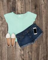Stylish summer outfit featuring a light blue top, denim shorts, and white slip-ons arranged on a rustic wooden background photo