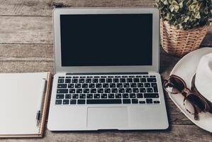 Workspace setup with laptop, notebook, sunglasses, and hat on wooden table photo