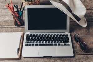 Workspace setup with laptop, notebook, hat, and sunglasses on wooden table photo