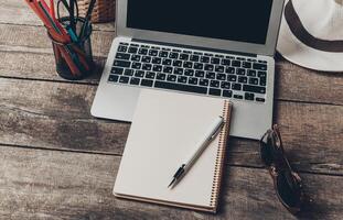 Work desk setup with laptop, notepad, pen, and sunglasses on a wooden table in a cozy environment photo