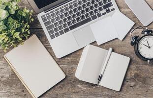 Workspace arrangement with laptop, notepad, stationery, clock, and greenery on wooden surface in a bright environment photo