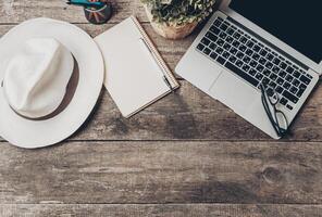 Workspace setup with a laptop, notebook, hat, and glasses on a rustic wooden table photo